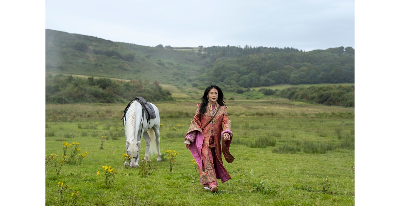 緑の草原のなかを、民族衣装姿の黒髪の女性が歩いている。女性の後ろに白い馬がいて、地面に頭を近づけている。遠くに森の木々。