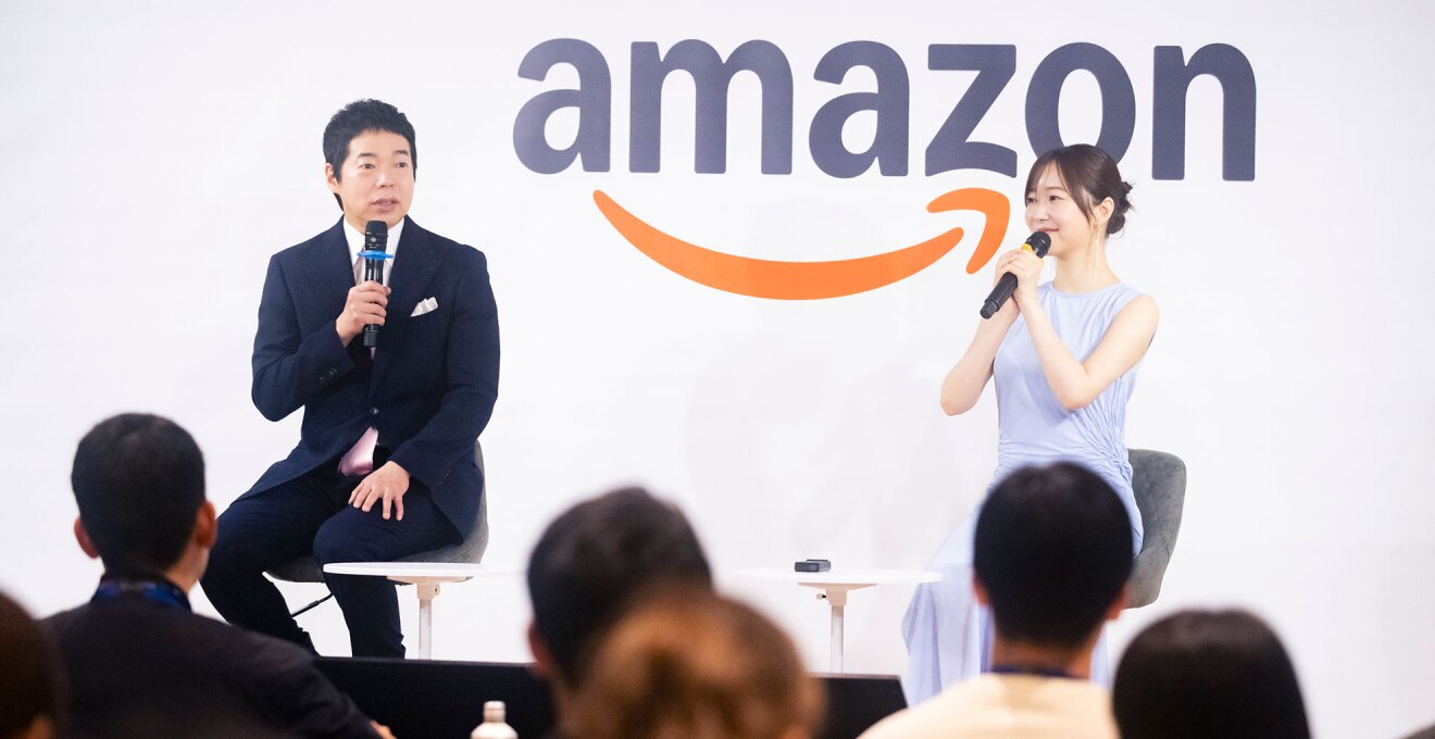 Amazonのロゴが大きく書かれた白い壁を背に、椅子に座った2人の人物。マイクを手に持って話をしている。手前に聴衆の後頭部が並ぶ。