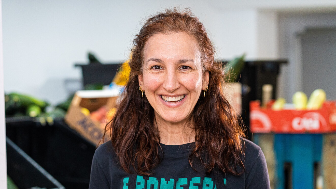 Amparo Zarza, Fundación Altius. LLeva un delantal de color negro y una camiseta negra de manga corta con las letras Promesas en verde. El pelo largo y castaño por el hombro, ondulado. Unos pendientes colgantes de oro. Mira a cámara y sonríe. De fondo se ven cajas de fruta- 