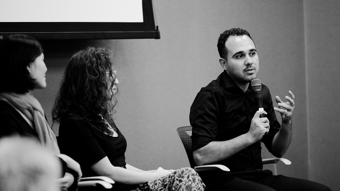 一名男子坐在椅子上,对着麦克风讲话,作为发言小组的一部分,左边是两名女性。