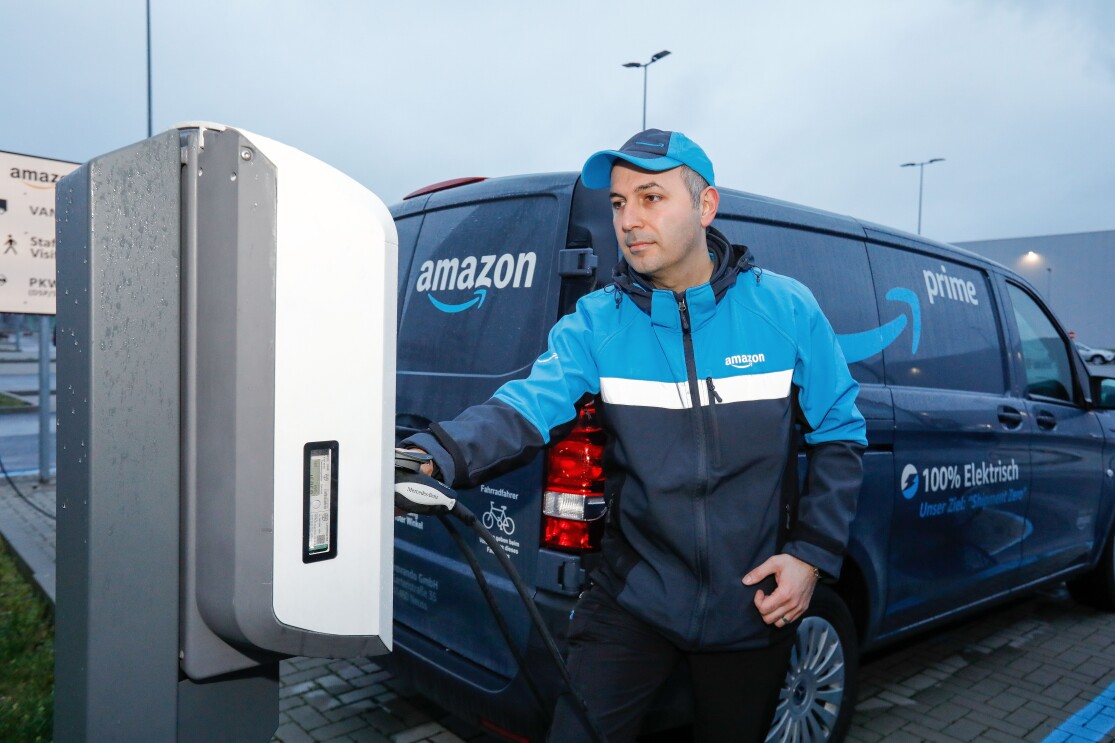 Ein Amazon Lieferfahrer an einer elektrischen Zapfsäule. Im Hintergrund sieht man das Lieferfahrzeug mit der Aufschrift "100 % Elektrisch"