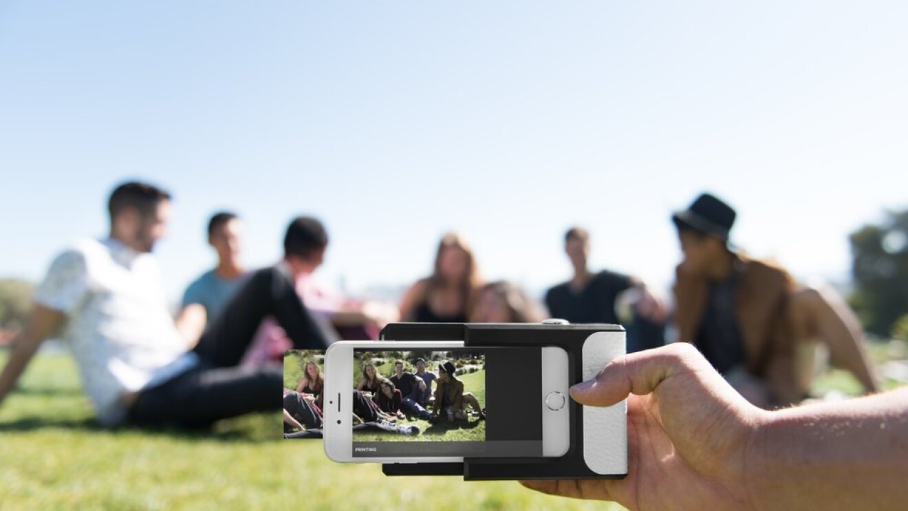 Une main tient une imprimante photo Prynt sur un smartphone, qui est en train d'imprimer la photo d'un groupe de jeunes gens assis dans l'herbe en extérieur