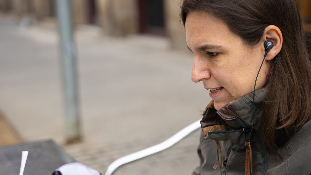 Gemma Viladomat, Senior Product Manager para Italia y España  de Amazon. Gemma trabajando en una terraza mientras toma un café. Una mesa plateada con un café y su cuchara. Ella está sentada mirandi fijamente a un ordenador que no se ve. Va con unos auriculares negros de cable y lleva la chaqueta puesta. Una cahqueta entre verde y gris y tiene el pelo castaño por el hombro. 