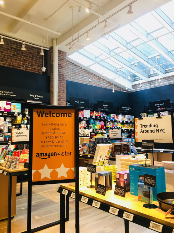 View inside Amazon 4-star. In the foreground, a sign that says "Welcome Everyting here is rated 4 stars & above, a top seller, or new & trending on Amazon.com. Next to the sign is a display table with products on it, and a "Trending Around NYC" sign. Some items on the table include Echo Show, Echo Dot, books and more. In the background, baby registry, baby and toddler products, grade school items, and more.
