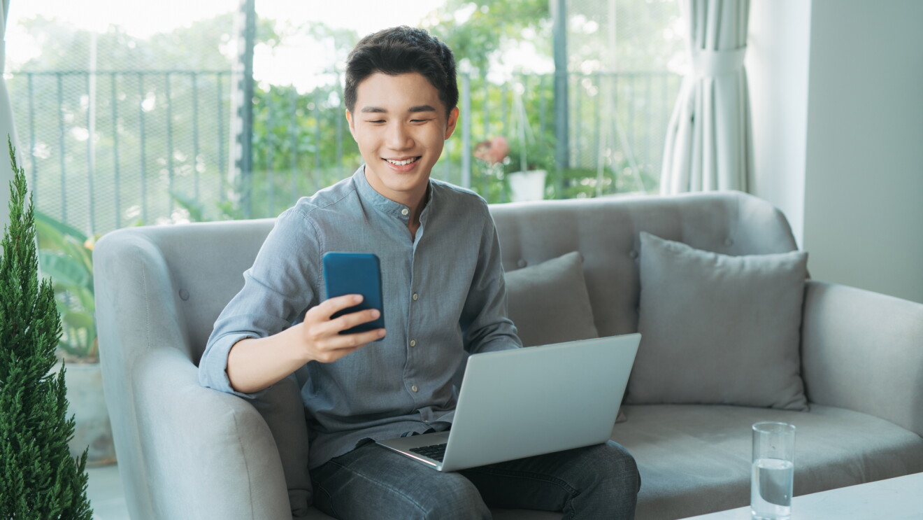 Young man smiling while using smartphone and laptop on couch

