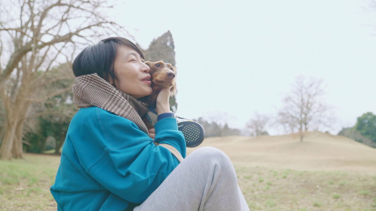 広い公園のような場所で愛犬のダックスフントにほおずりしている植村さん