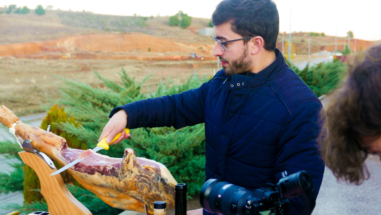 David González, CEO de Iberic Box cortando jamón. Momento de la grabación del vídeo en el que la cámara aparece en el margen inferior derecho. En primer plano, David González, cortando un jamón que está en un jamonero en una mesa situada en el exterior. El fondo son campos. 