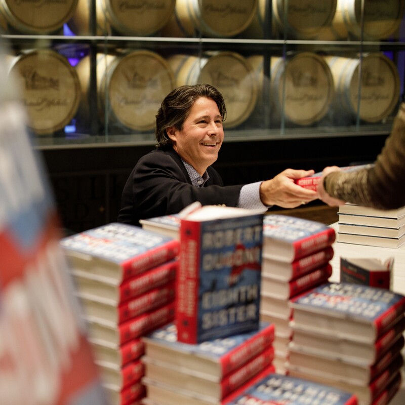 A man sits at a table covered with stacked copies of books. He hands a book another person.