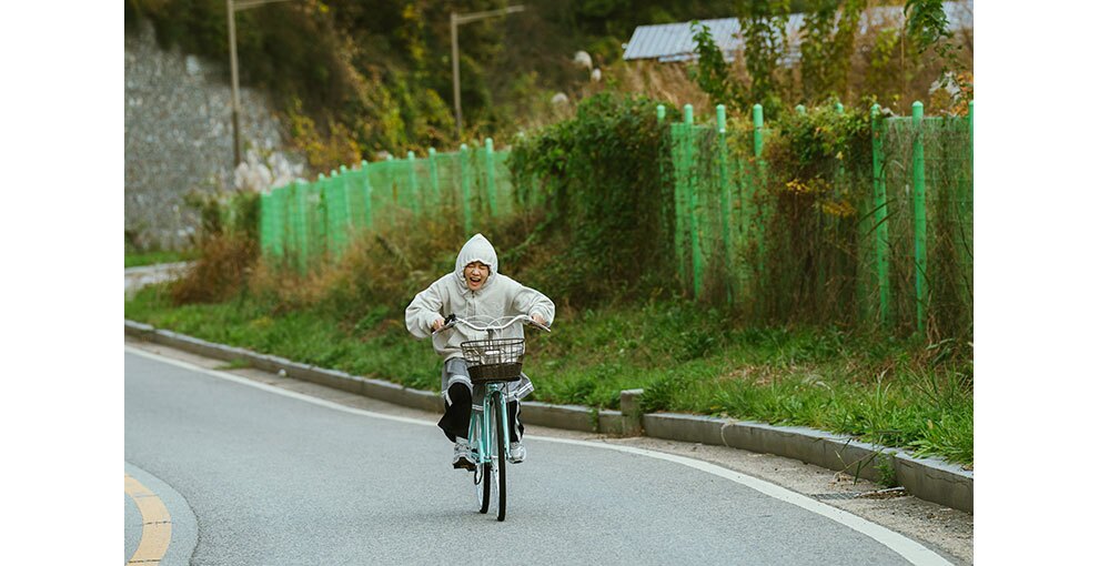 自転車をこいで舗装道路を走る人物。着用したパーカーのフードをかぶっている。道路の右側にはフェンスと背の高い植物やツタ。