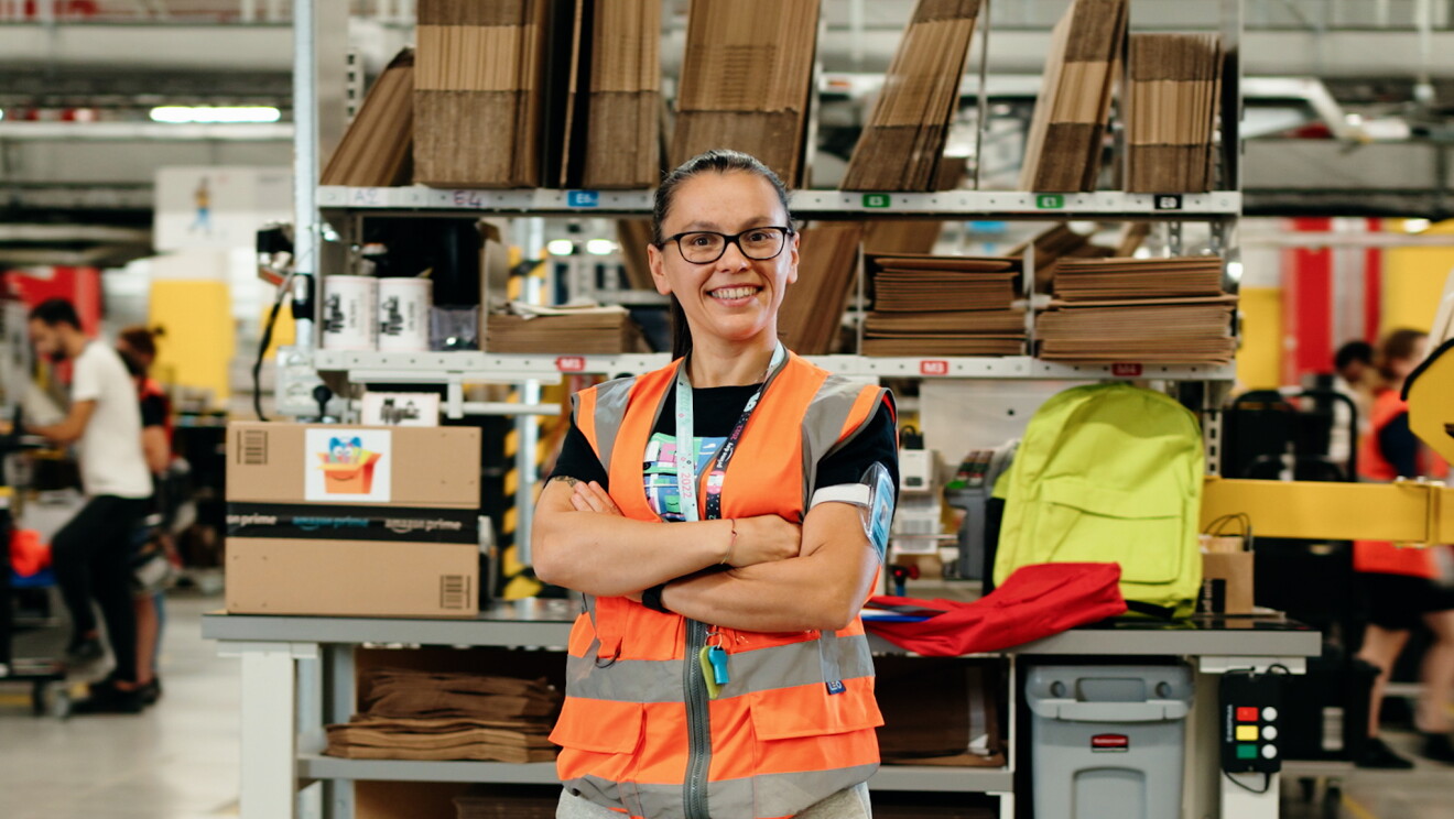 Samantha Pavesi, trabajadora de Amazon en el centro logístico de Novara (Italia). Aparece en primer plano con los brazos cruzados y de pie mirando a cámara. Con una sonrisa y unas gafas de color negro. Lleva un chaleco de color naranja. De fondo se ve una mesa de empaquetar de un centro logísticos y por los márgenes personas difuminadas. 