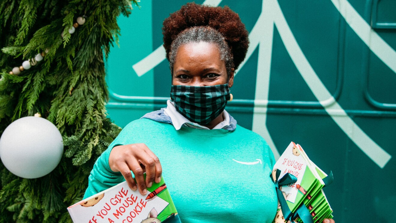 An image of a woman smiling for a photo while wearing a mask. She is holding stack of children's books and wearing a green Amazon shirt. Behind her is a large wreath with ornaments on it.