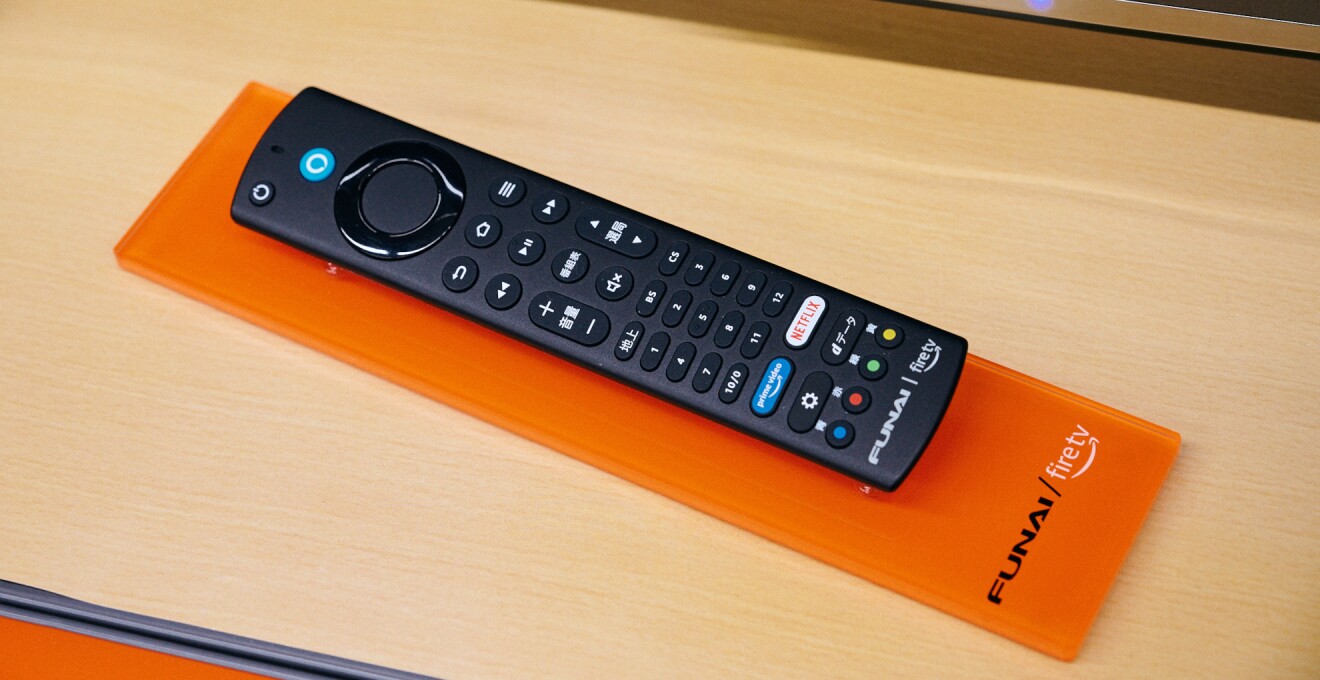 黒い「FUNAI Fire TV搭載スマートテレビ」のリモコン。テレビのリモコンに似ているが、少し細長く、先端近くに青い丸のAlexaのボタンがついている