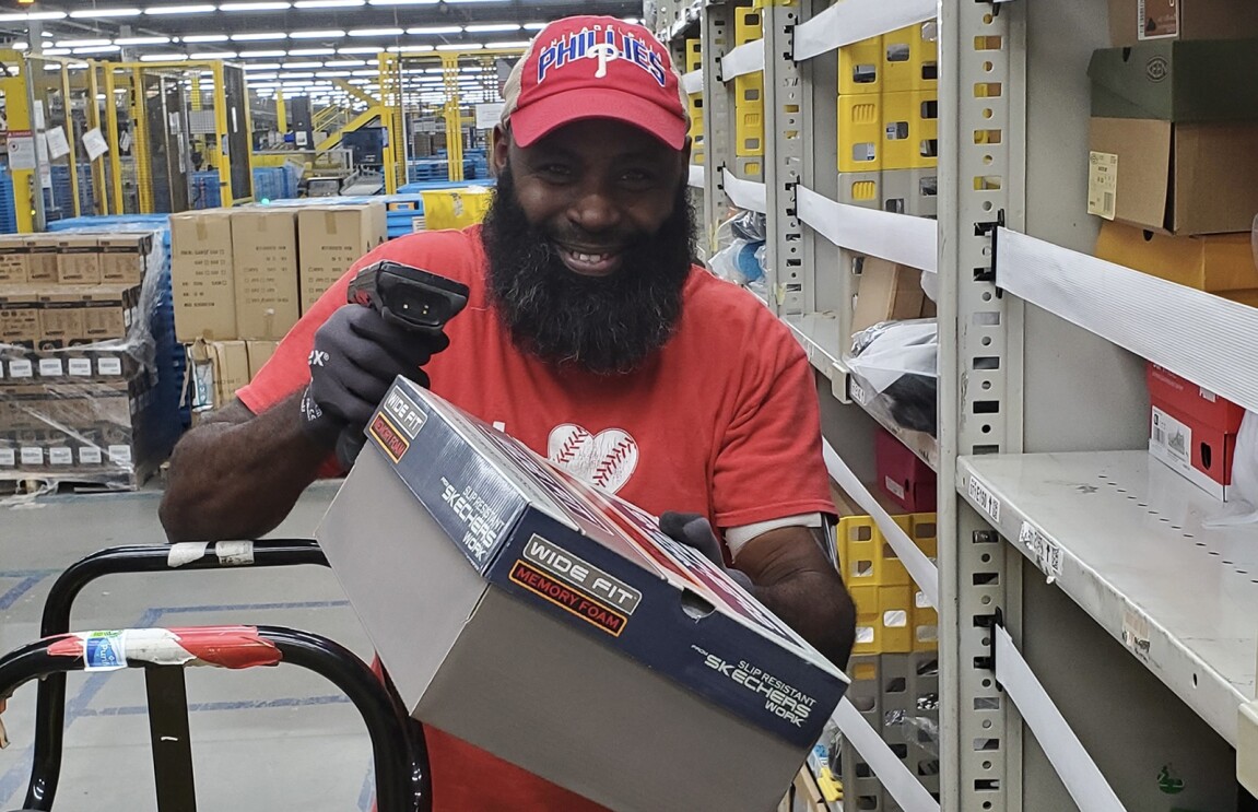 An image of a man in an Amazon fulfillment center smiling while holding a box of shoes in one hand and a bar code scanner in the other.