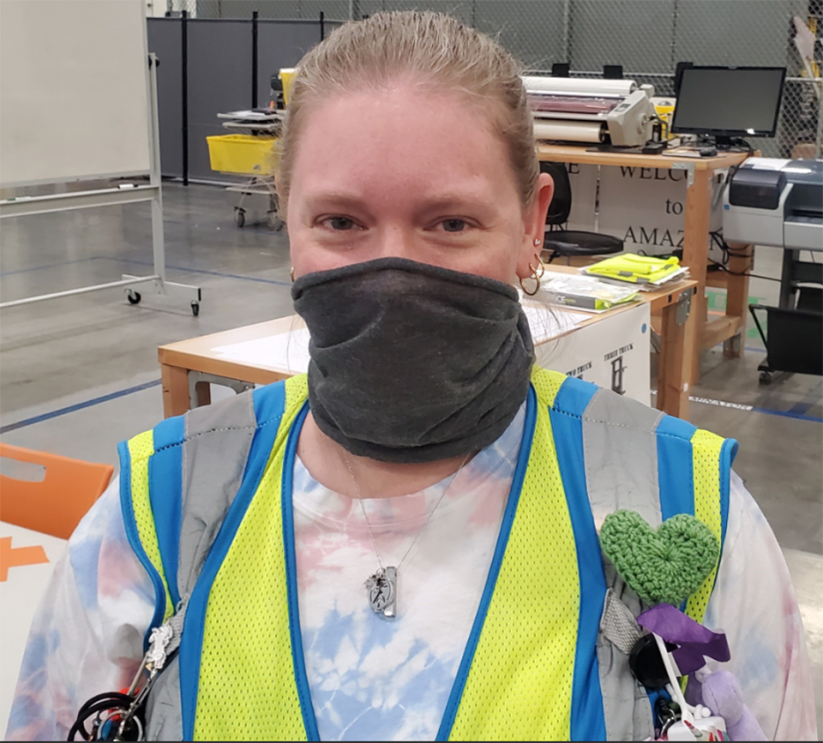 A woman wears a face mask and safety vest. On her vest, a green, crocheted heart pin is attached. 