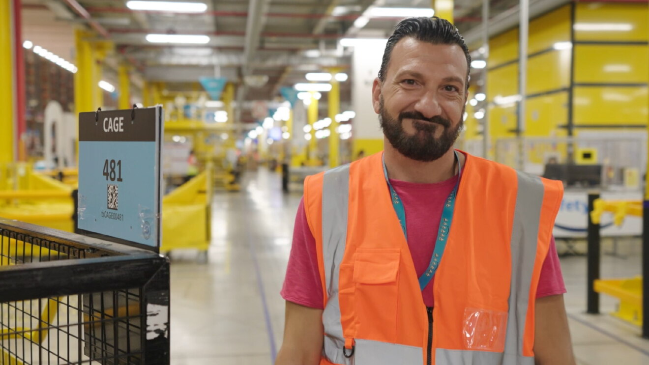 Mezzo busto di un dipendente Amazon che sorride verso la camera. Indossa un giubbino di sicurezza arancione