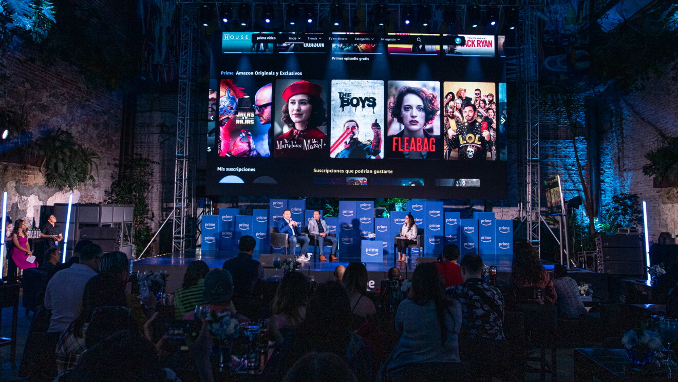 En un escenario con cajas con el logo de Prime, aparecn tres personas sentadas que no se pueden identificar y encima de ellas los cárteles de las series y películas más conocidas en México. 