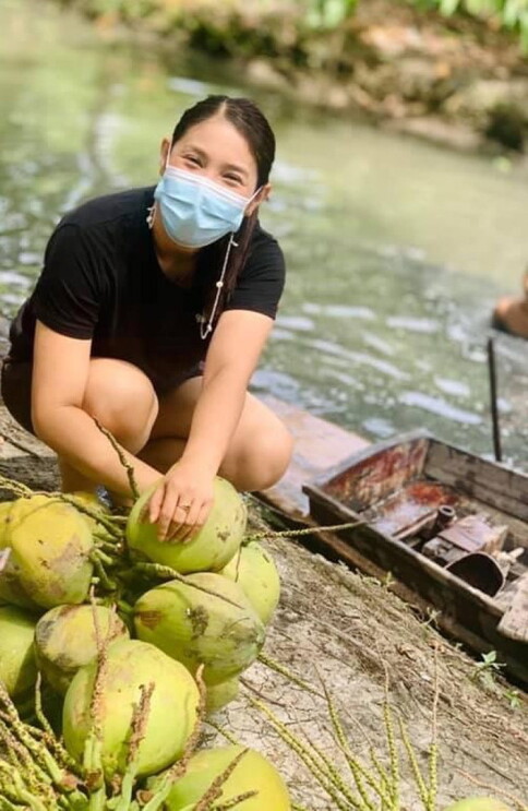 An image of a woman working near a river with a coconut in front of her. She has a surgical mask on.