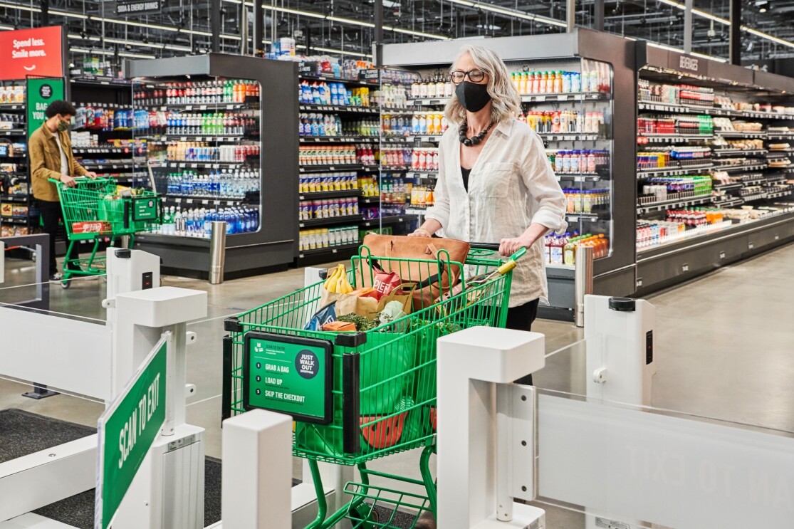 A woman with short curled blonde hair pushes her green grocery cart filled with bags of grocery and her purse through the exit of the Just Walk Out gates.