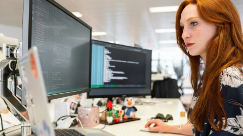 Una donna con i capelli rossi seduta ad una scrivania mentre lavora al computer. Di fronte a sè ha due schermi e un laptop.
