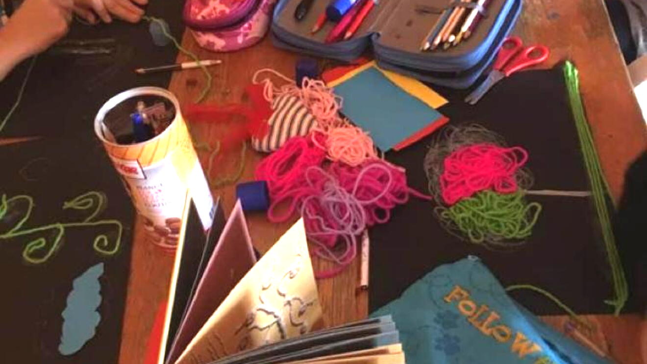 Primo piano di un tavolo su cui sono poggiati astucci, libri, matasse di cotone e colori. Sui lati si intravedono le mani di alcuni bambini che stanno colorando su degli album da disegno. 