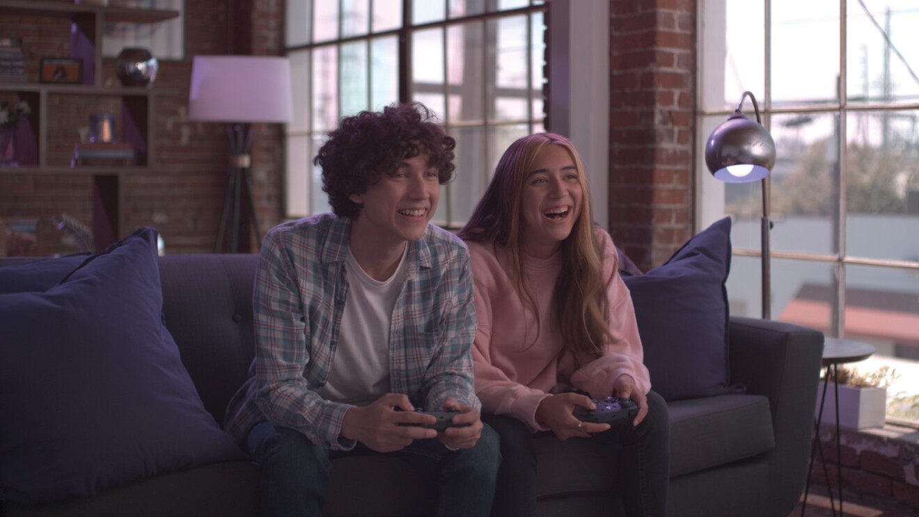 A young man and young woman sitting on a couch in a city apartment holding Luna controllers and playing video games and having fun