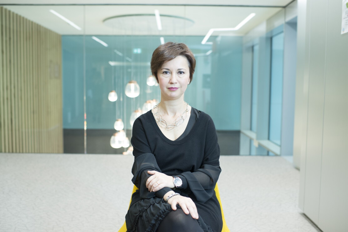 Mariangela Marseglia, directora General de Amazon España e Italia. En un espacio con una silla de color amarillo y un armario color blanco, aparece sentada en esa silla Mariangela Marseglia. Con el pelo corto de color castaño, tiene los brazos cruzados, e una muñeca tiene un reloj y en la otra una pulsera. Va con un vestido negro por la rodilla y encima un jersey negro de manga corta y de punto. Tiene un collar de piezas ovaladas unidas. De fondo se ve un cristal y una lámpara colgando del techo y que parece que da a un hall amplio. Si bien una de las paredes es un armario, la otra son unos tableros verticales de madera. 