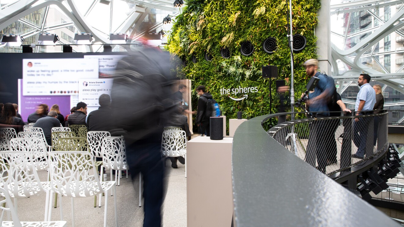 A man walking into the September 2018 Devices announcement event. To the left are chairs in front of a large screen. To the right others stand against a railing. Behind the scene is a living wall in The Spheres.