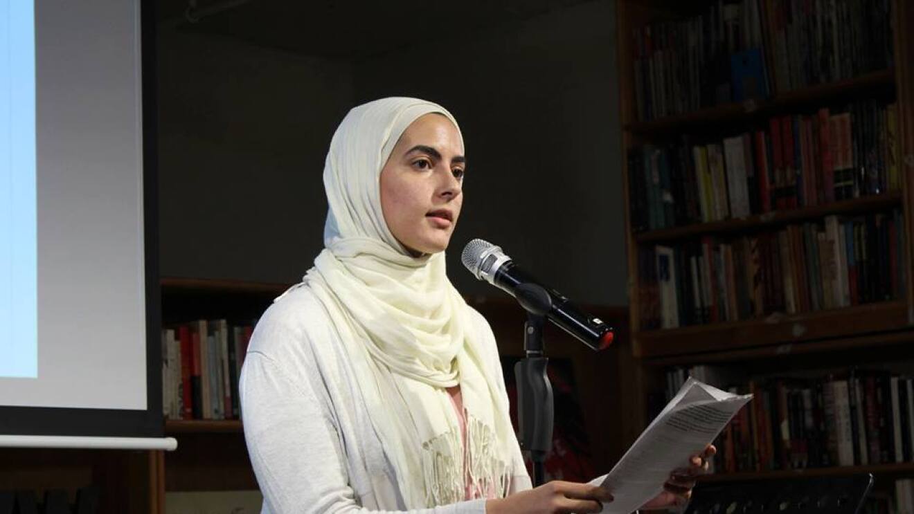 A woman (Sumaya Awad) stands in front of a microphone, speaking about her experience