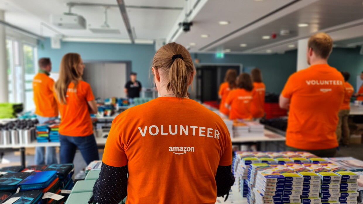 Man sieht Amazon Mitarbeiter:innen mit orangenem Volunteer-T-Shirt von hinten. Sie stehen hinter Tischen mit Schulmaterialien.