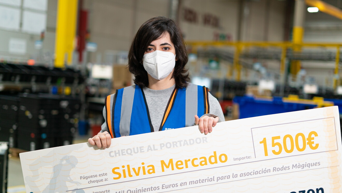 Silvia Mercado es la cuarta ganadora de una Amazon Dream Box en España. Silvia está sujetando un cheque de 1 500 euros para material para la asociación Rodes màgiques. De fondo hay un centro logístico con estanterías de color amarillo. Silvia lleva el pelo  negro por el hombro y escalonado. Su jerseiy es de color gris y lleva un chaleco de Amazon Box Star de color azul. 