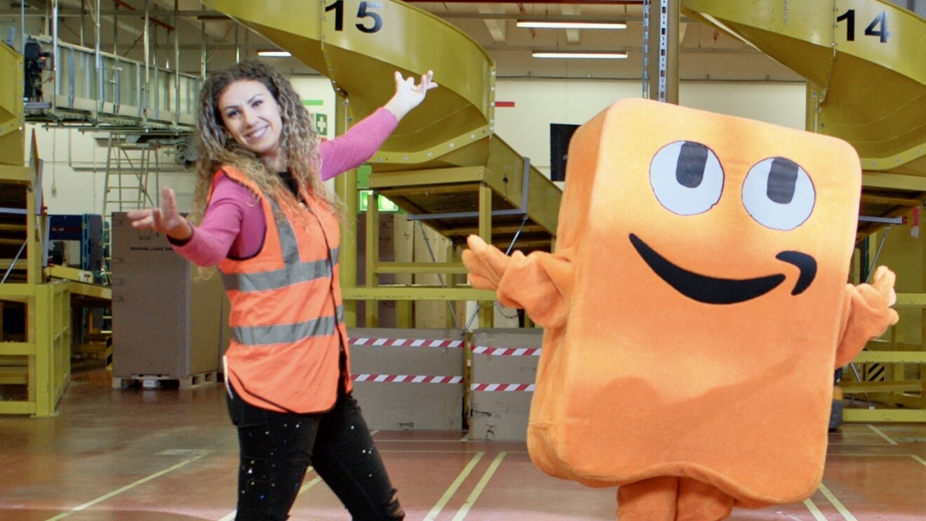 Eine blonde Amazon Mitarbeiterin neben dem Amazon Maskottchen Peccy.