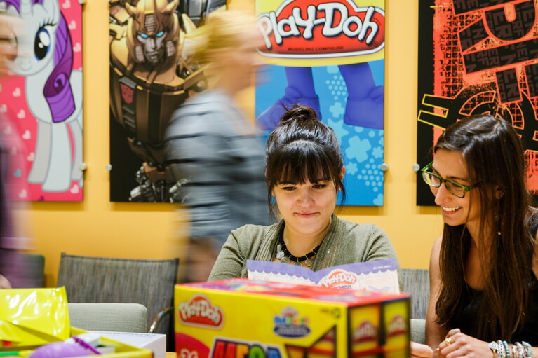 Two women in the foreground sit at a table surrounded by packaging. Other people pass by in the background. The wall has posters of My Little Pony, Play-Doh, and other Hasbro brands.