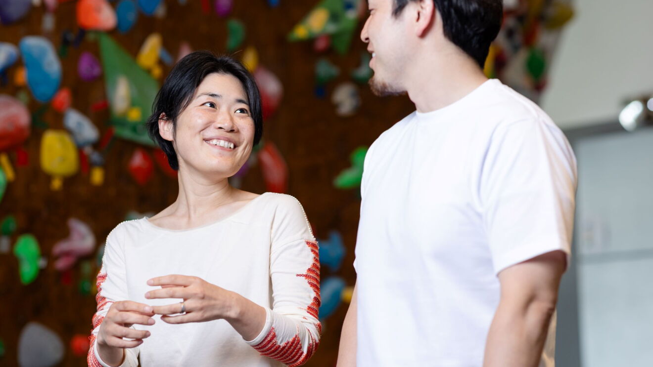 短髪の女性が微笑みながら隣に立っている白いTシャツを着ている男性を見ている。彼らの後ろにはカラフルなボルダリングウォールが見える。