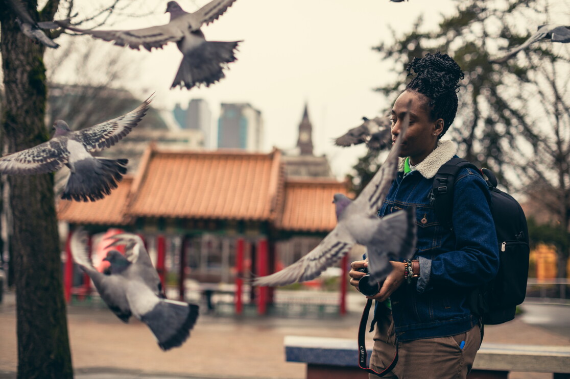 A woman in a blue jacket, green top, and brown pants, and holding a camera, walks in front of a temple. Flying birds are in the foreground. 