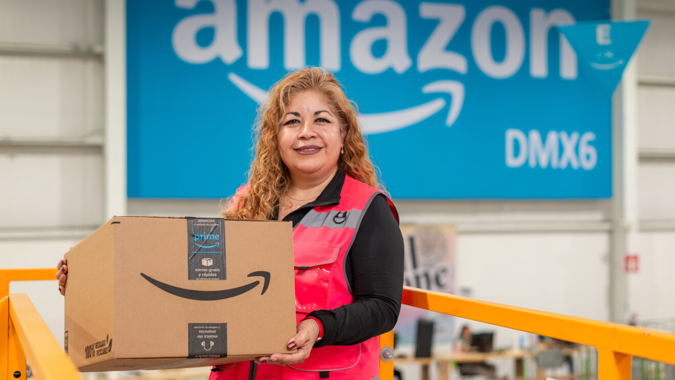 Trabajadora de Amazon cargando una caja delante de un poster que pone Amazon DMX6.