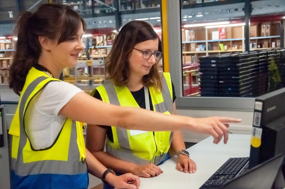 Isabelle steht links neben Larissa, im Hintergrund Regale mit Wein und Spirituosen. Isabelle hält einen Scanner in der Hand, Larissa eine Likörflasche.