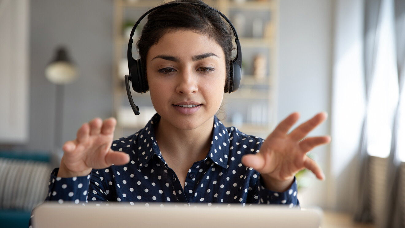 A woman wearing a button down shirt and headset looks into her laptop screen while gesturing with her hands. 
