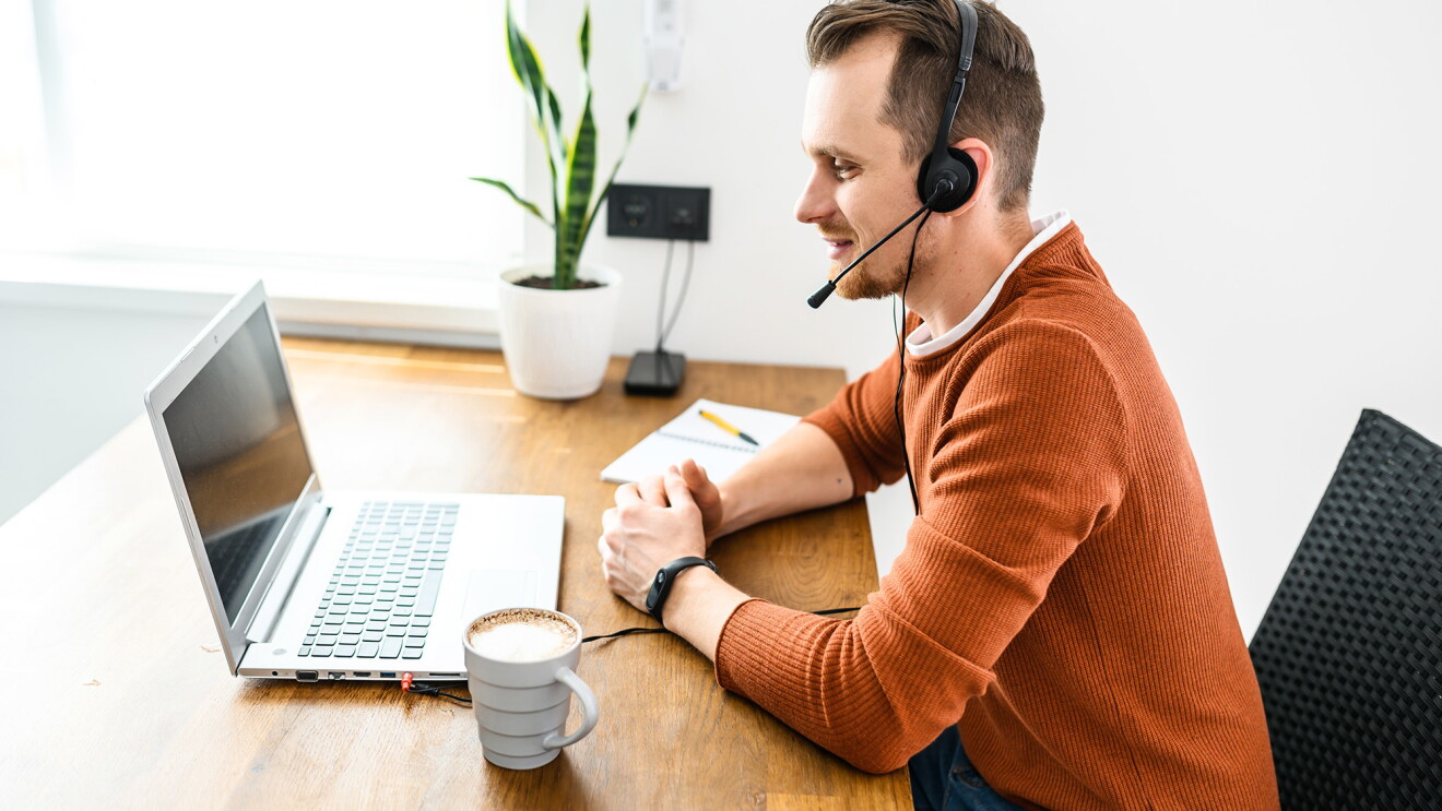 A man wears a headset and works on a laptop computer, with a notepad and coffee mug nearby. 