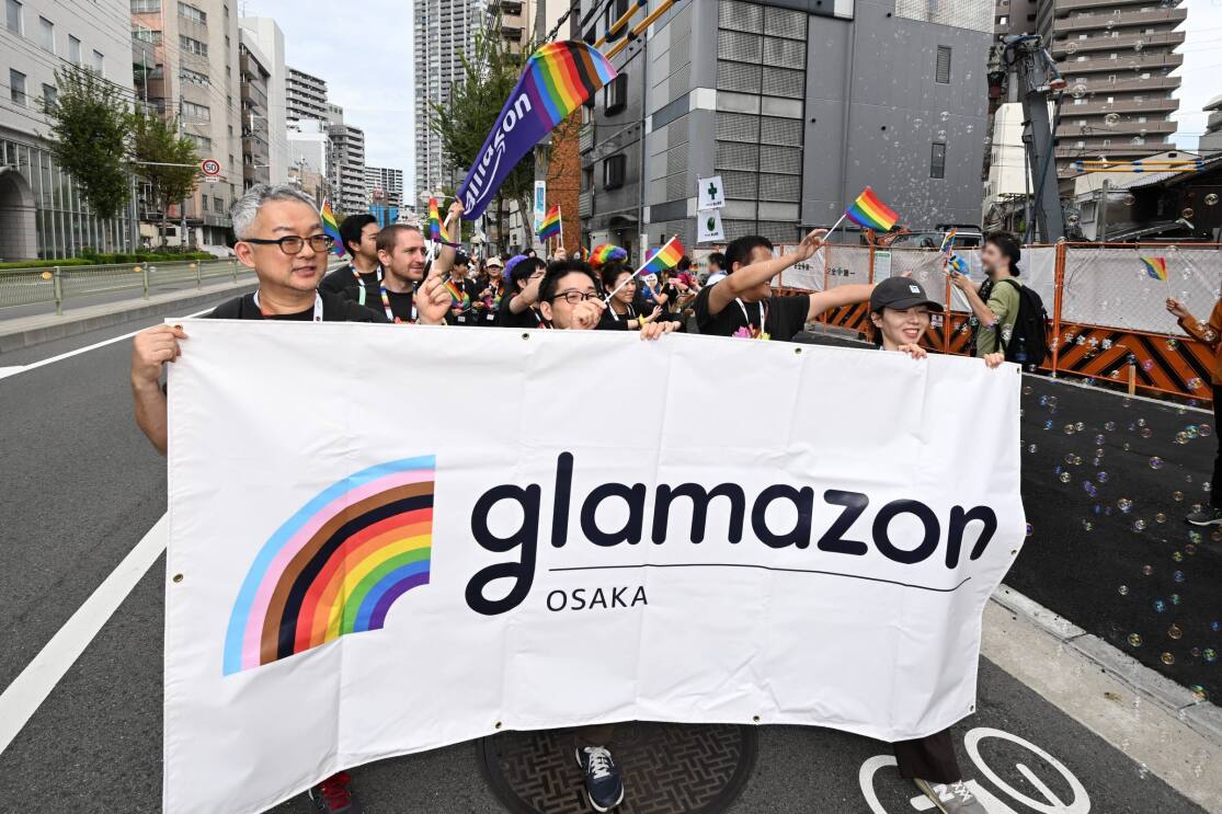 メガネをかけている男性2人と帽子を被っている女性がGlamazon Osakaの横断幕を持って、グループの先頭を歩いている。彼らの後ろには数人の人々が続いている。そのグループの右側にはたくさんのシャボン玉が飛んでいる。