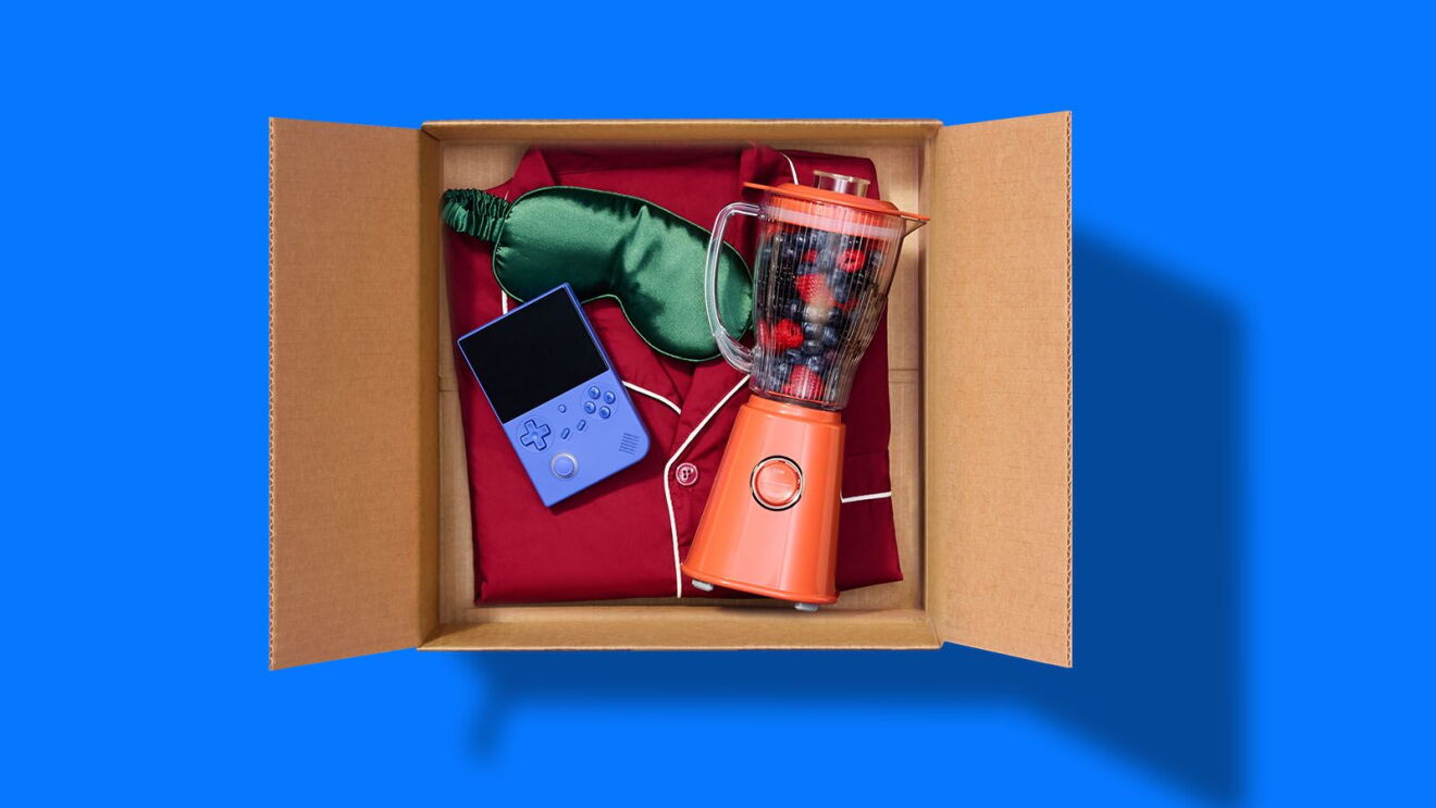 A cardboard box containing red pajamas, a green sleep mask, a blue handheld game console, and an orange blender filled with berries