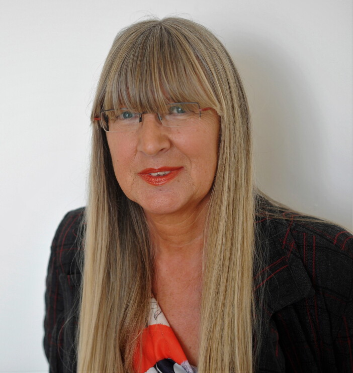 Eine Frau mit blonden langen Harren und Brille lächelt in die Kamera. Sie trägt roten LIppenstift.