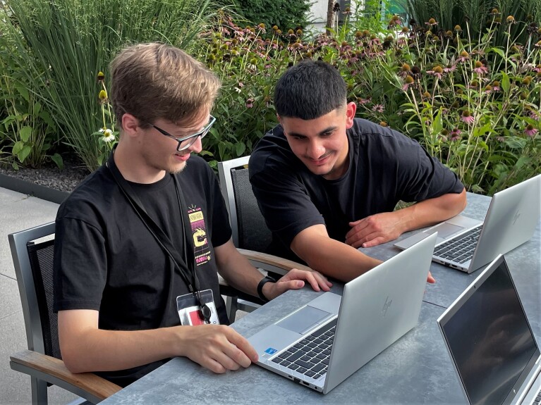 Die zwei AWS Azubis Hendrik und Mohammed sitzen draußen vor ihren Laptops und lächeln.