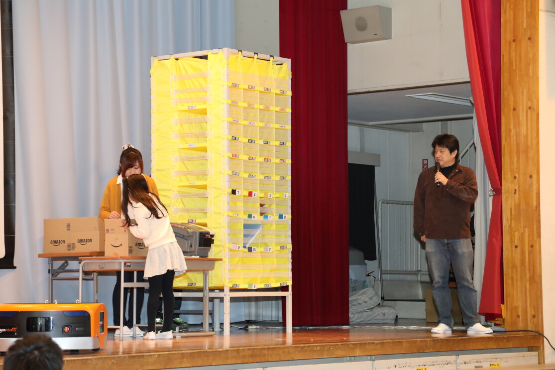 体育館のステージ上に設置された模擬倉庫で作業をする子供。子供の前には机を並べた台があり、ダンボール箱がのっている。右側に黄色い商品分類棚。ステージ右端にマイクを持って立つ男性。