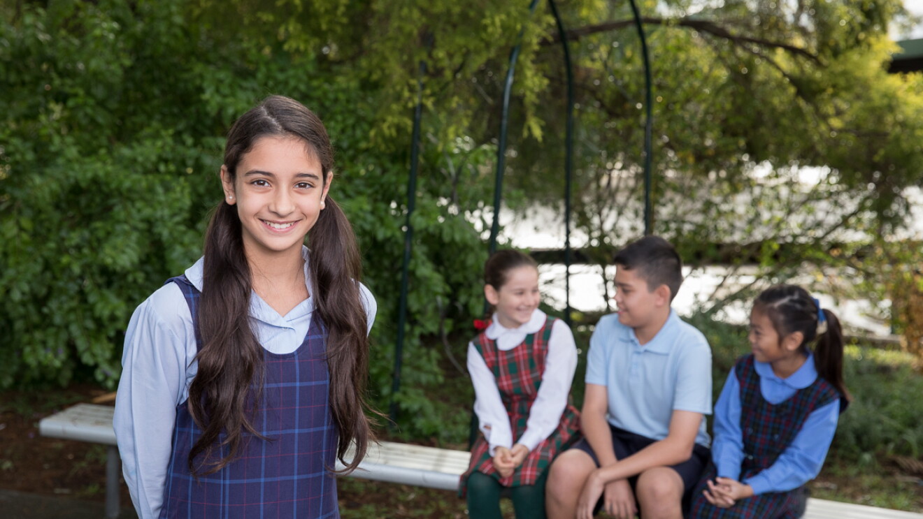 A young girl smiles and poses for the camera while three of her classmates sit behind her smiling at one another. They are outside surrounded by greenery and wear school uniforms.