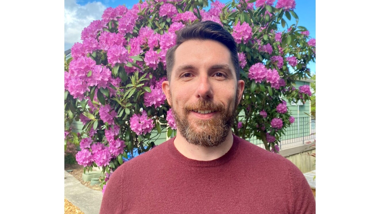 Fabio smiling, wears a red long-sleeved shirt has a beard. He is standing in front of a tree with purple flowers.