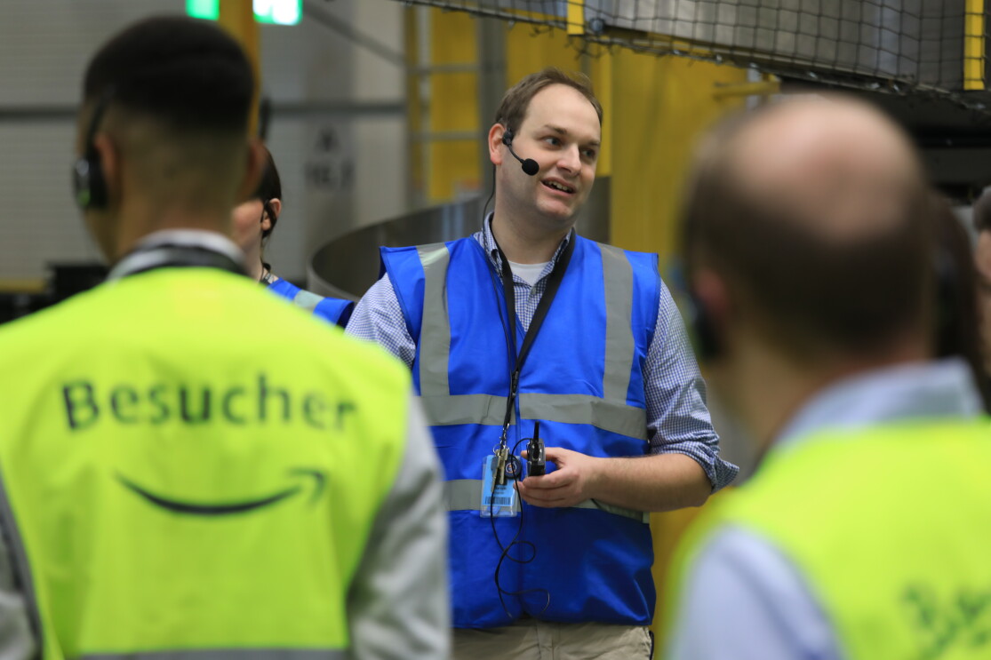 Ein Tourguide in blauer Sicherheitsweste steht vor Besuchern, die eine gelbe Sicherheitsweste tragen. 