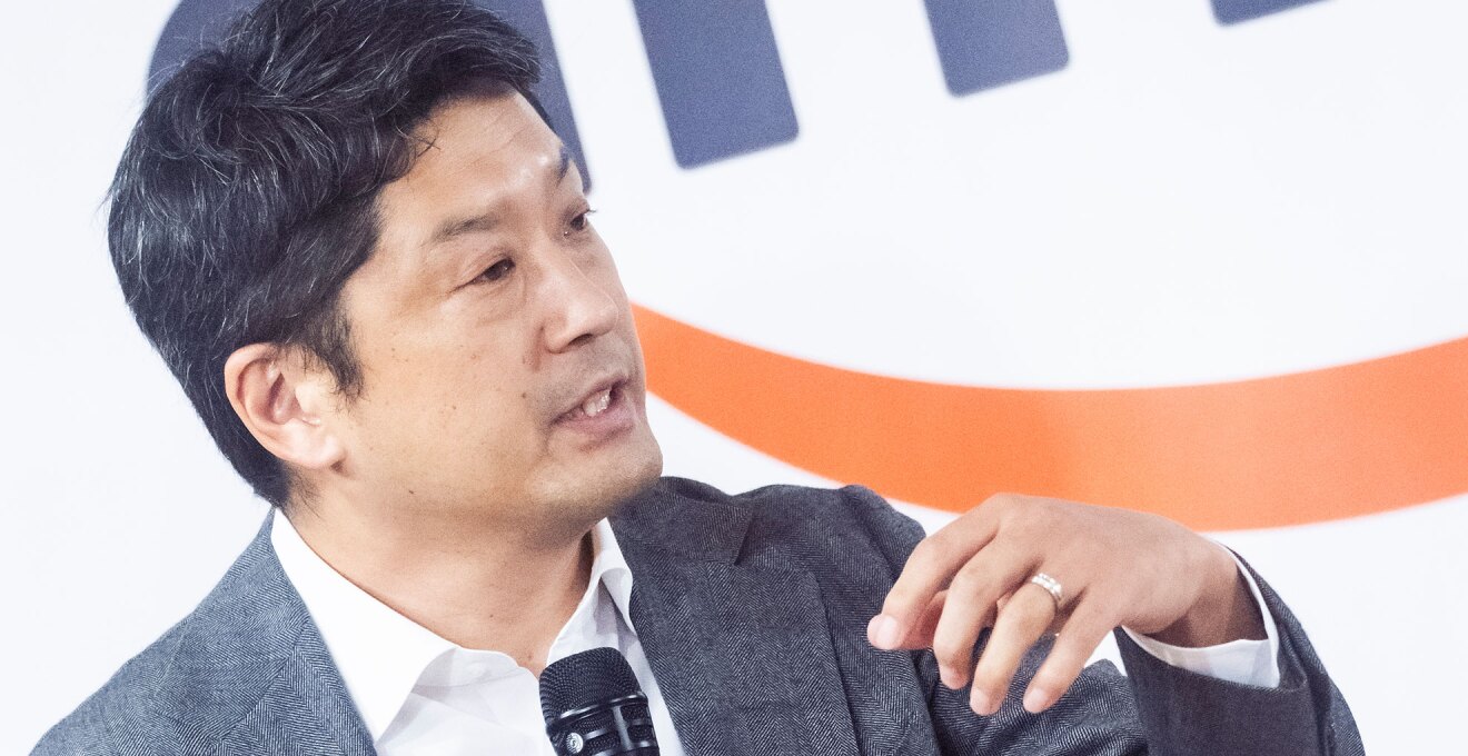 身振りを交えながらマイクに向かって話をする男性。グレーのスーツ着用でネクタイはなし。背後の壁にオレンジの模様が見える。