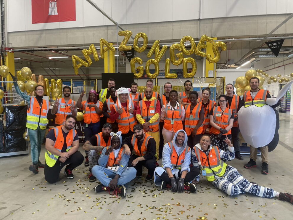 Eine große Gruppe von Amazon Mitarbeiter:innen steht und liegt vor dem goldenen Schriftzug „Amazon Goes Gold“.