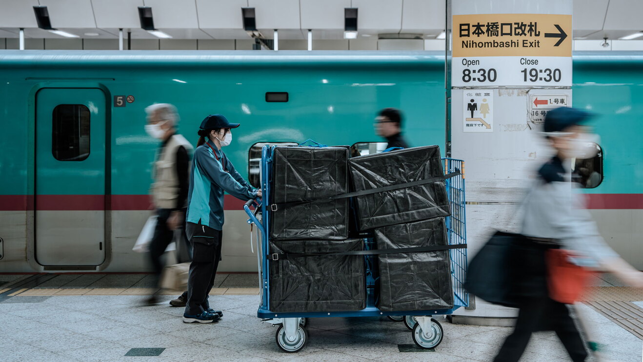 新幹線が停車中のプラットフォームを、ボックスが4つ積まれた台車を押して歩く作業員。ホームを歩く他の人はぼやけている。