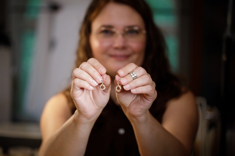 A woman smiles as she holds a pair of earrings towards the camera.